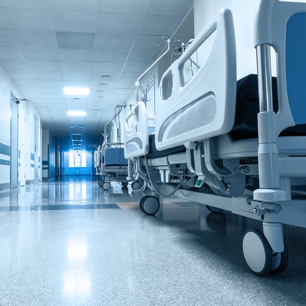 Empty hospital beds lined up in a clean, brightly lit hallway with reflective floors, white walls, and doors leading to patient rooms.