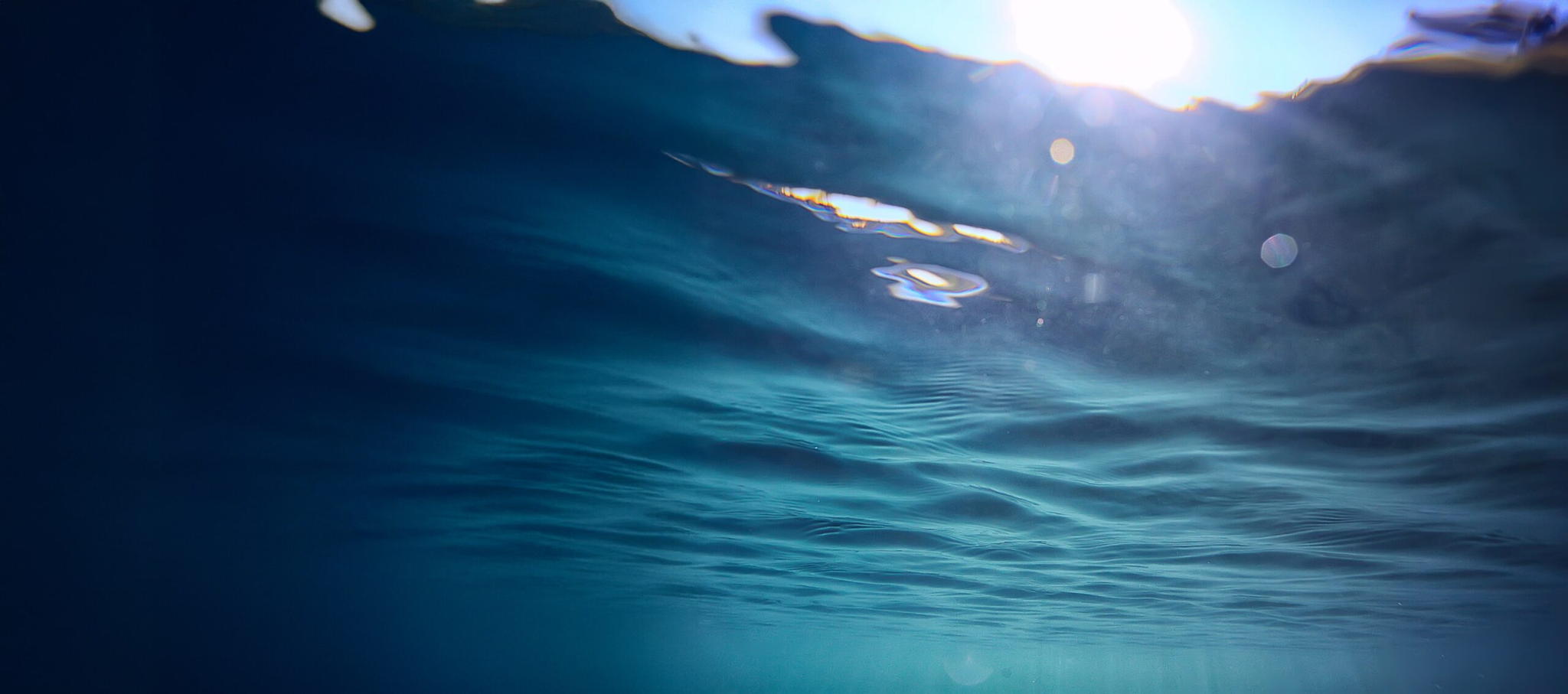 An underwater view looking up at the sun shining through the rippling surface of deep blue water, creating patterns of light and shadow.