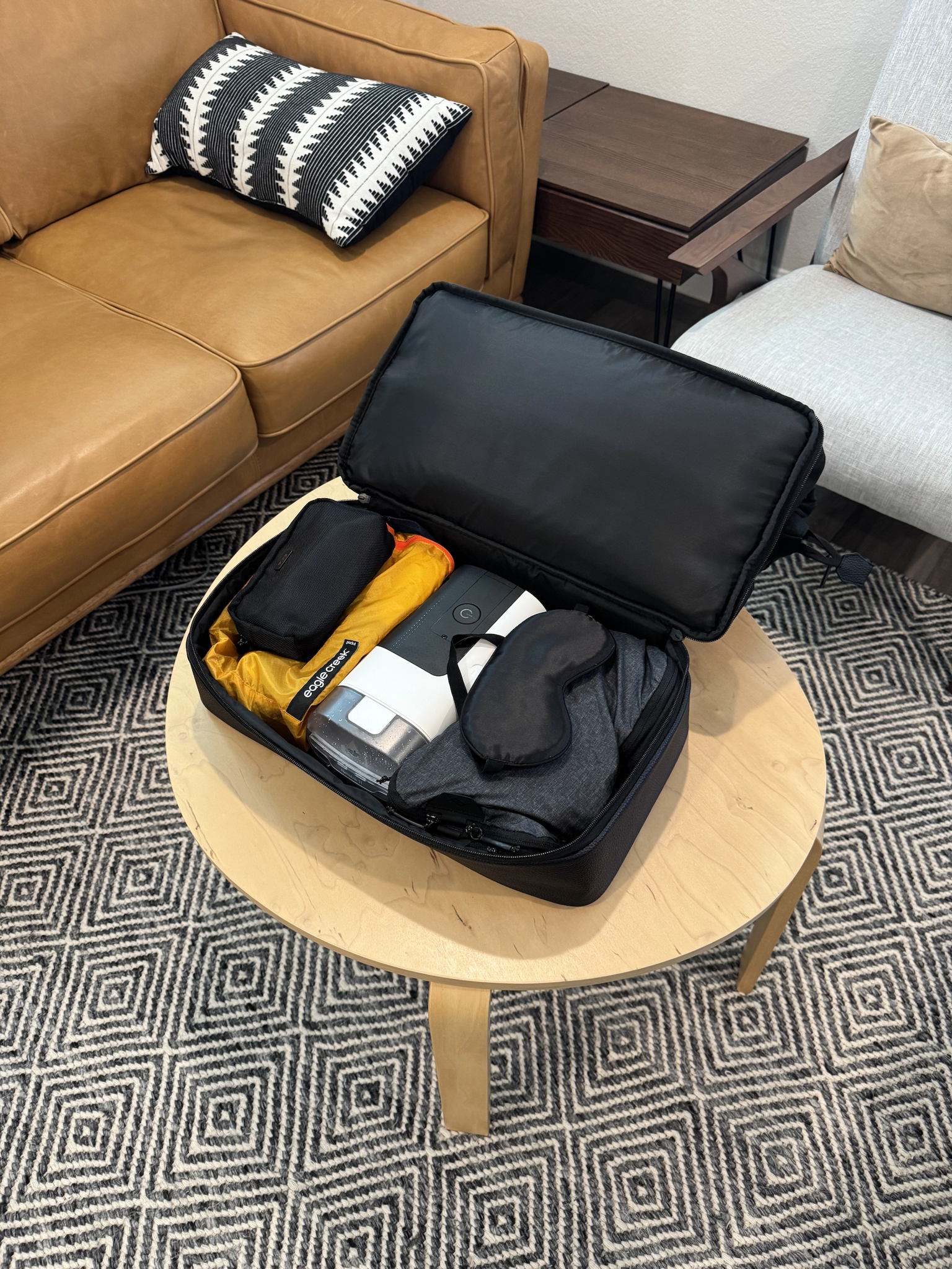 Weekender bag with zippered bottom compartment open to reveal a CPAP machine and neatly folded clothing inside. The bag sits on a tidy bed in a softly lit room, creating an organized and calm atmosphere. No visible text in the image.