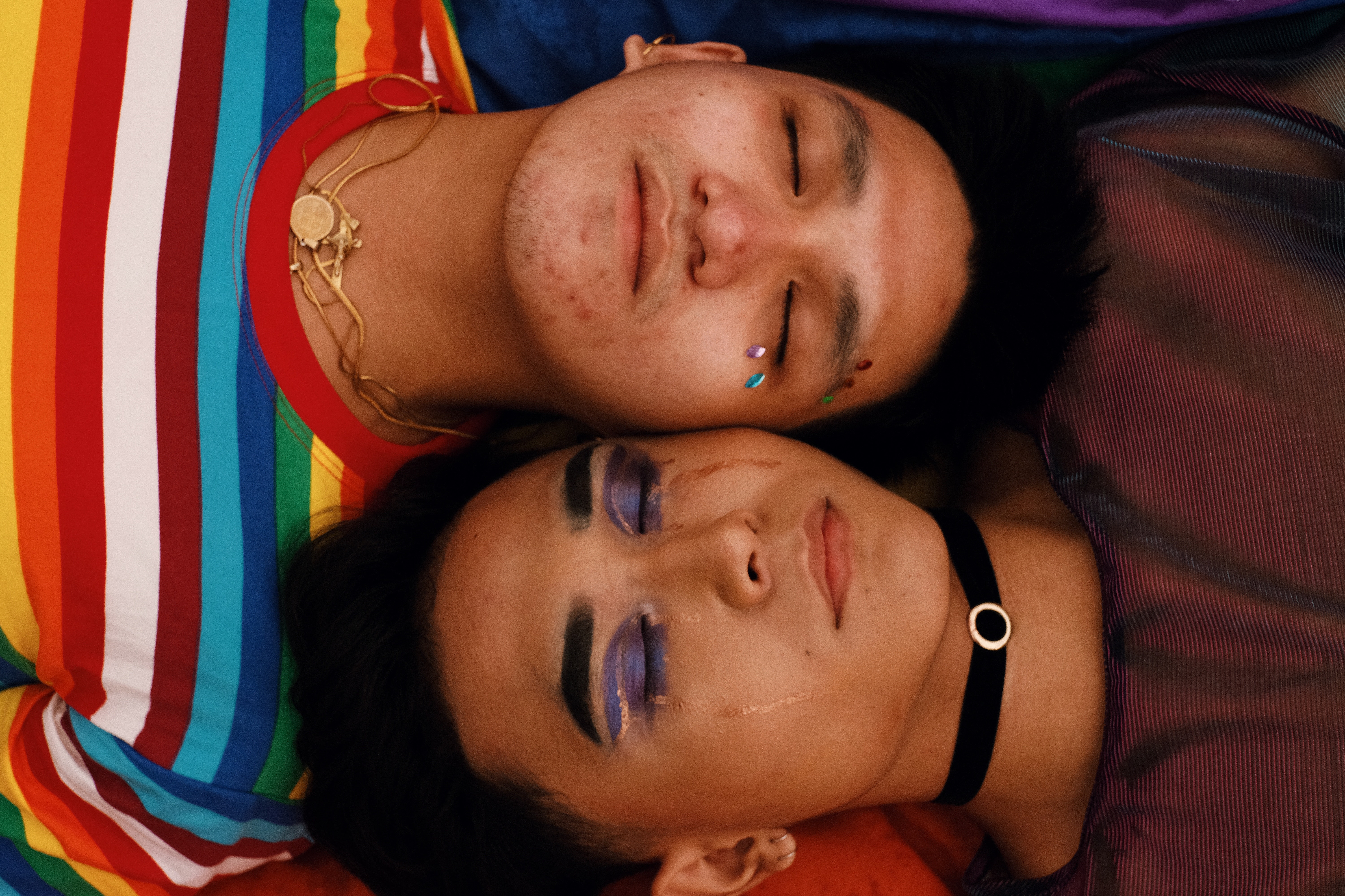 Two bi racial friends lay in the floor with their heads touching and their eyes closed wearing colorful clothing.