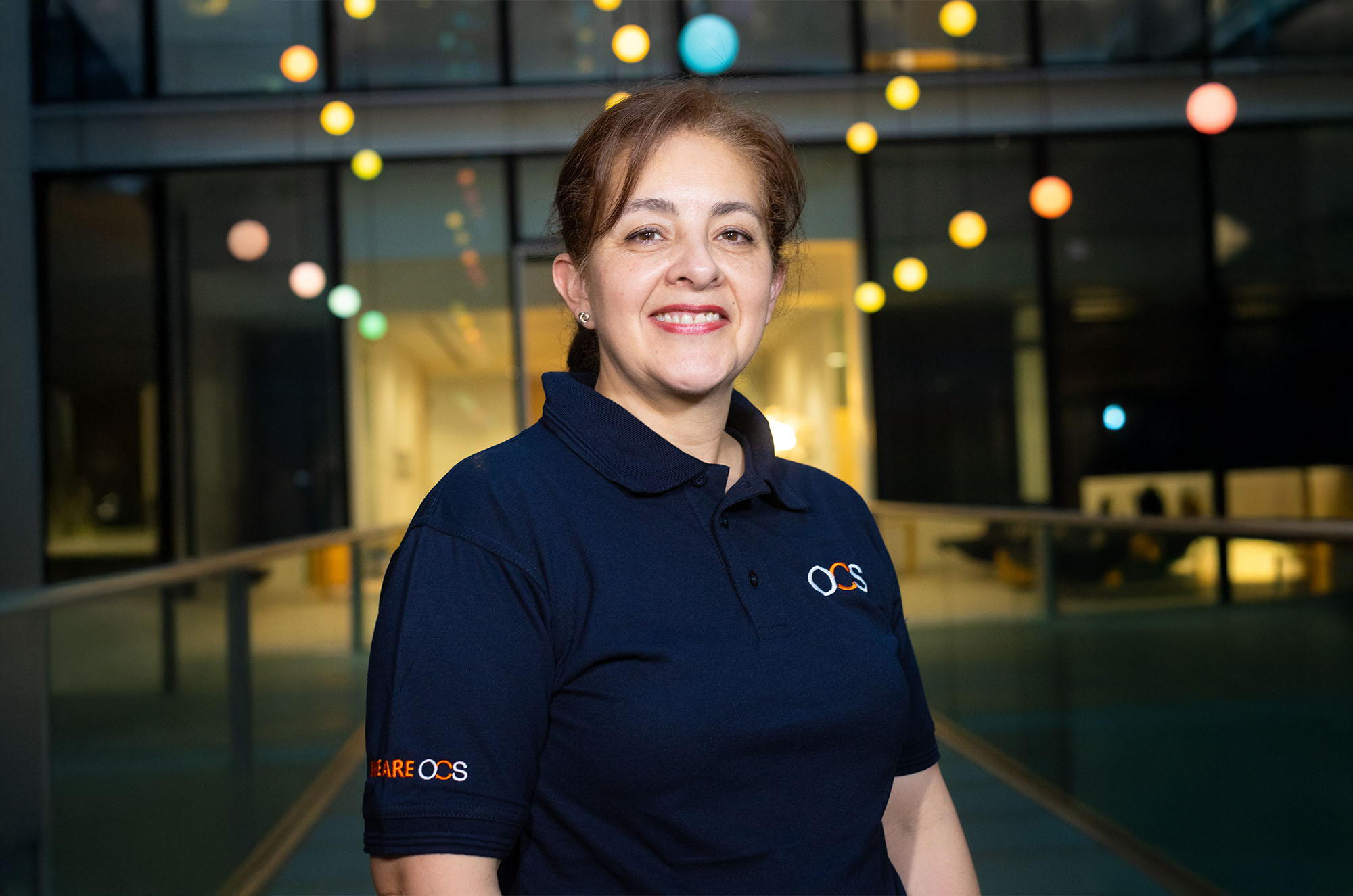 A woman with brown hair tied back, wearing a navy blue polo shirt with logos, stands smiling indoors in front of glass walls with colorful hanging lights in the background.