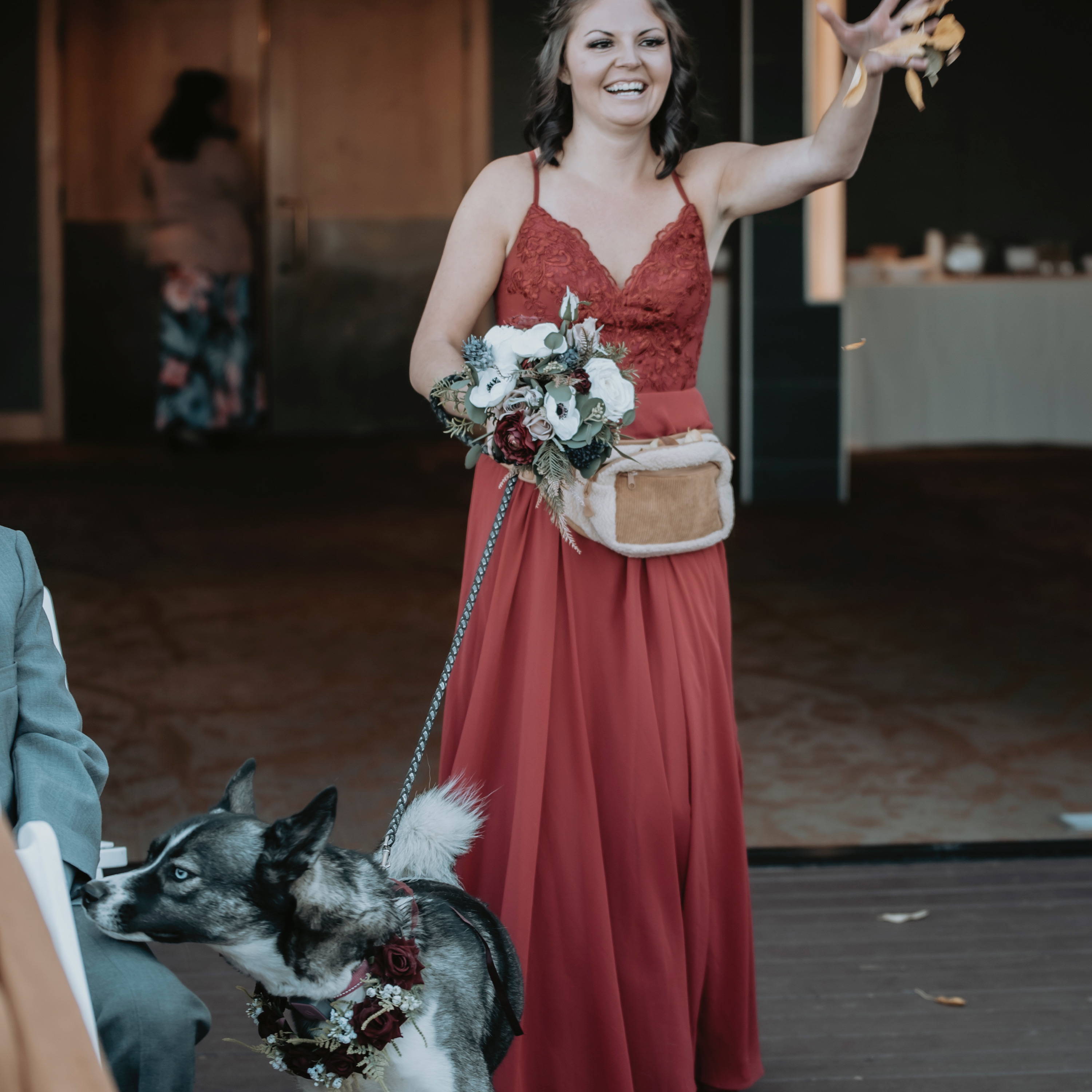 A bridesmaid wearing a terracotta dress sprinkles rose petals while walking a dog wearing a floral collar down the aisle for a wedding ceremony 