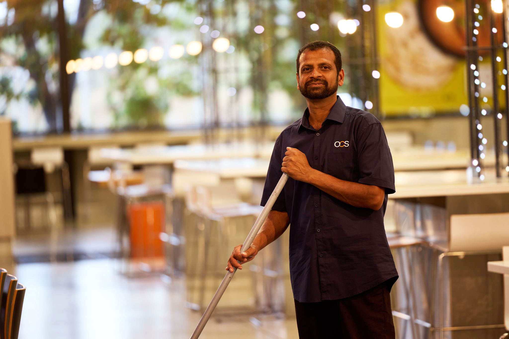 A man in a navy blue uniform is mopping the floor in a bright, modern, and empty cafeteria. He is looking at the camera and smiling slightly.