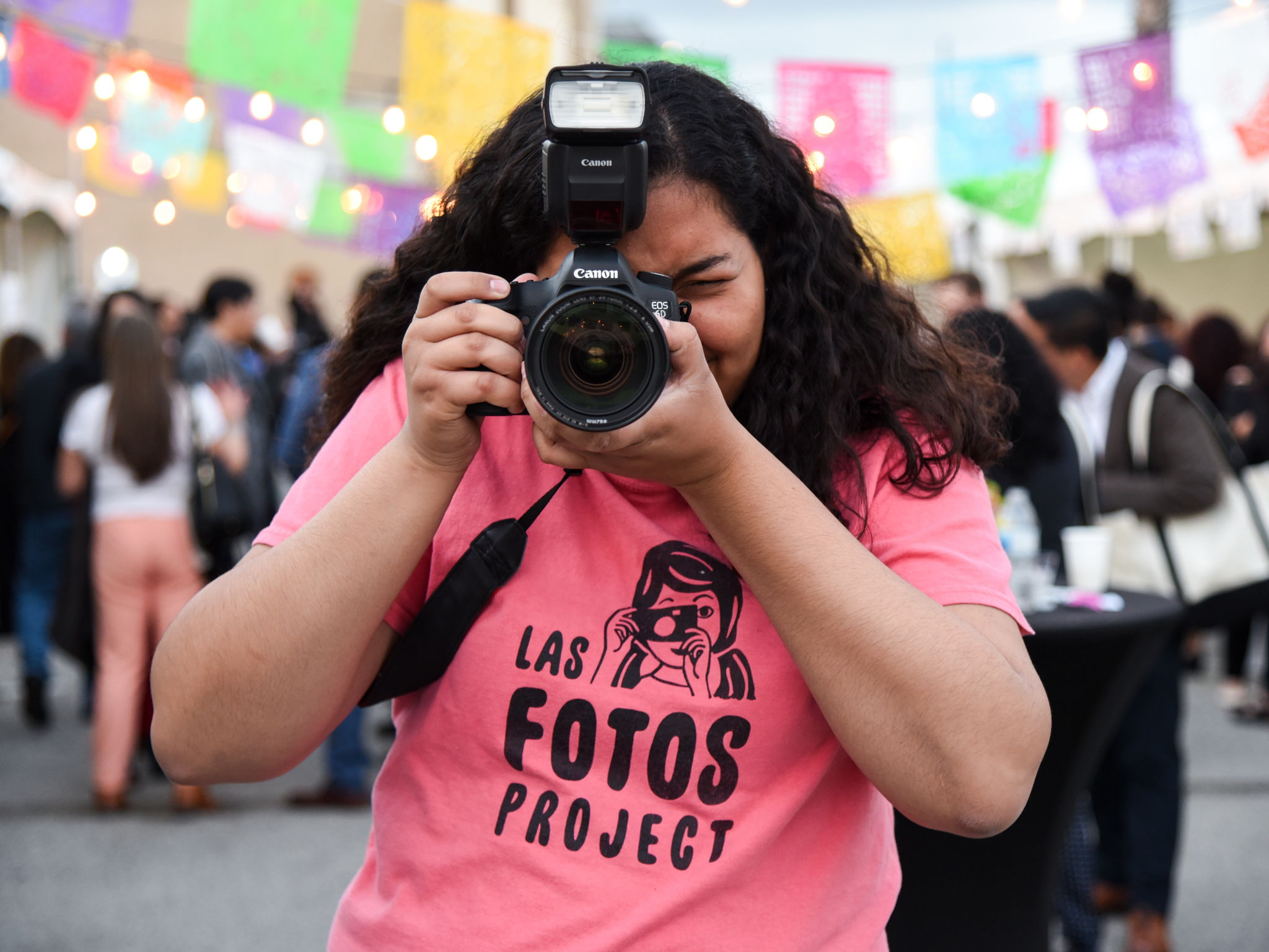 A girl wearing a pink "Las Fotos Project" t-shirt holds a DSLR camera up to her face