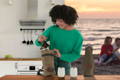 woman putting a growler in a growler pack with a kitchen and beach behind her