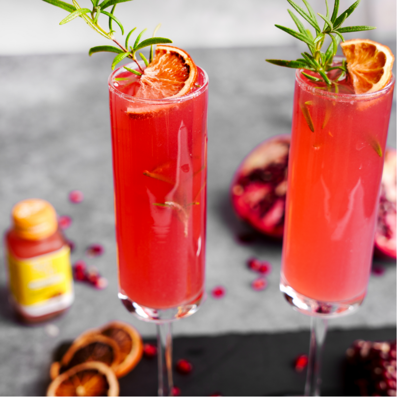 two glasses of a red Mimosa Mocktail with a slice of orange and a Bottle of IMMUNI-TI SuperShot 