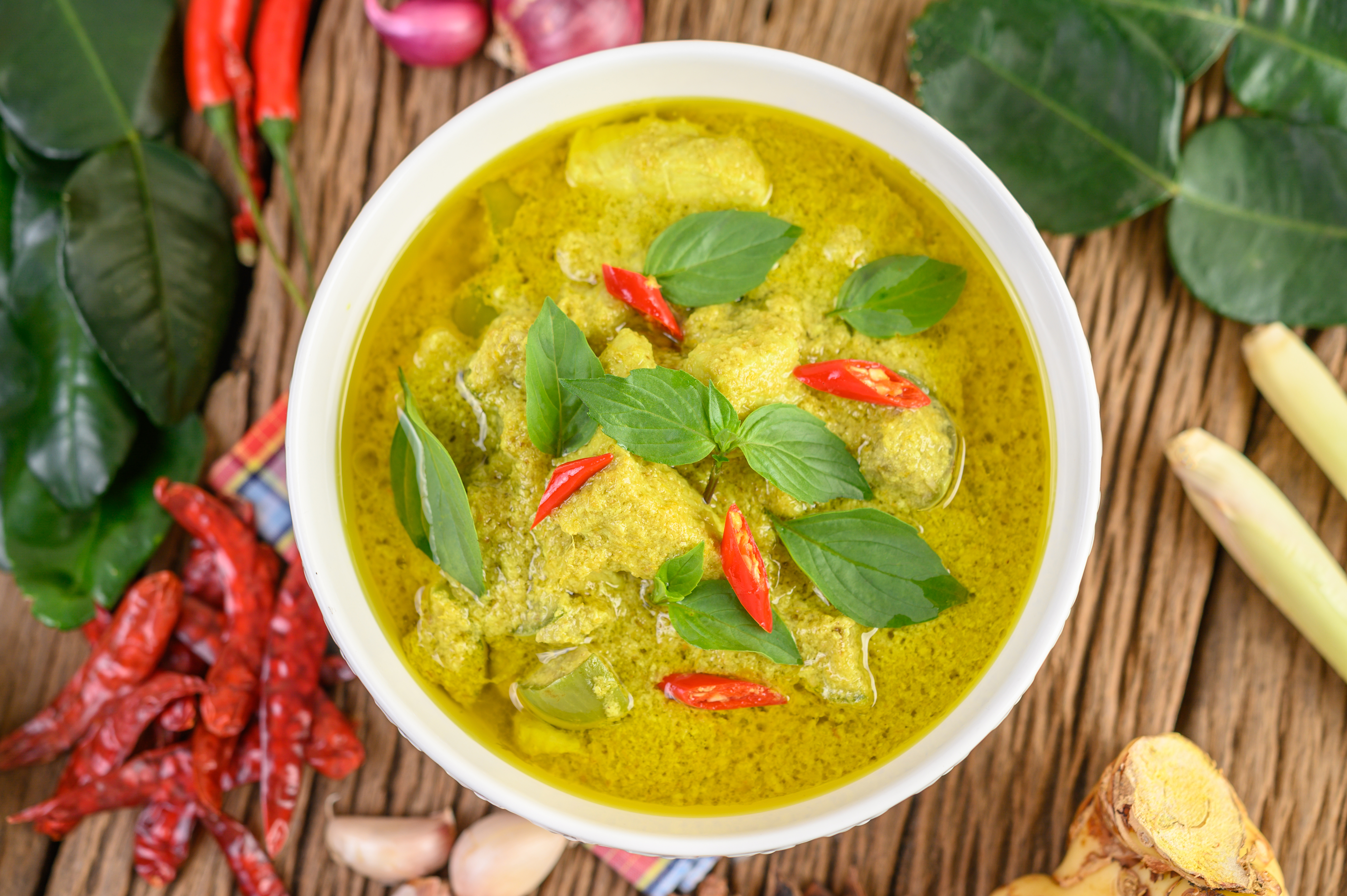 A bowl of green curry with basil and chiles on top surrounded by fresh curry ingredients