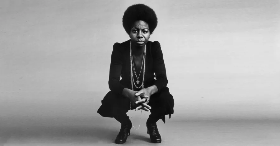 Nina Simone crouched down with her hands together, wearing black and her hair in an afro.