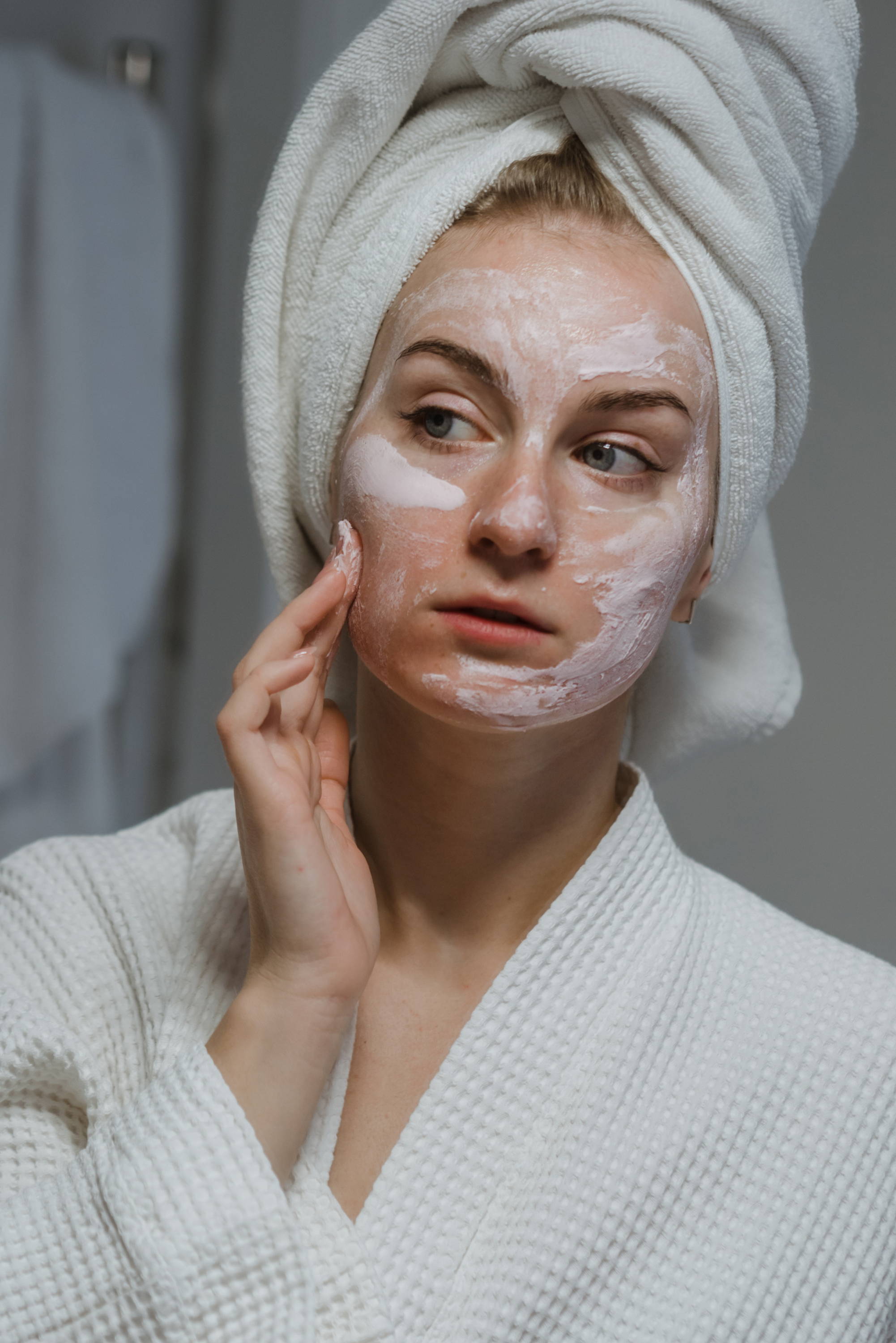 woman applying a face mask to her skin with her hair wrapped in a towel whilst looking in a mirror