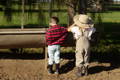 Two children in farm clothes and rubber boots standing beside an animal feeding trough outdoors.