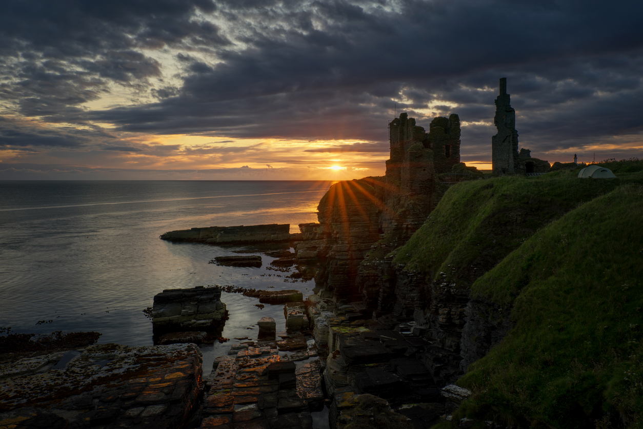 The sun sets over the ruins of a stone castle on a grassy cliff by the sea, casting golden light across the water and illuminating the dramatic clouds in the sky.