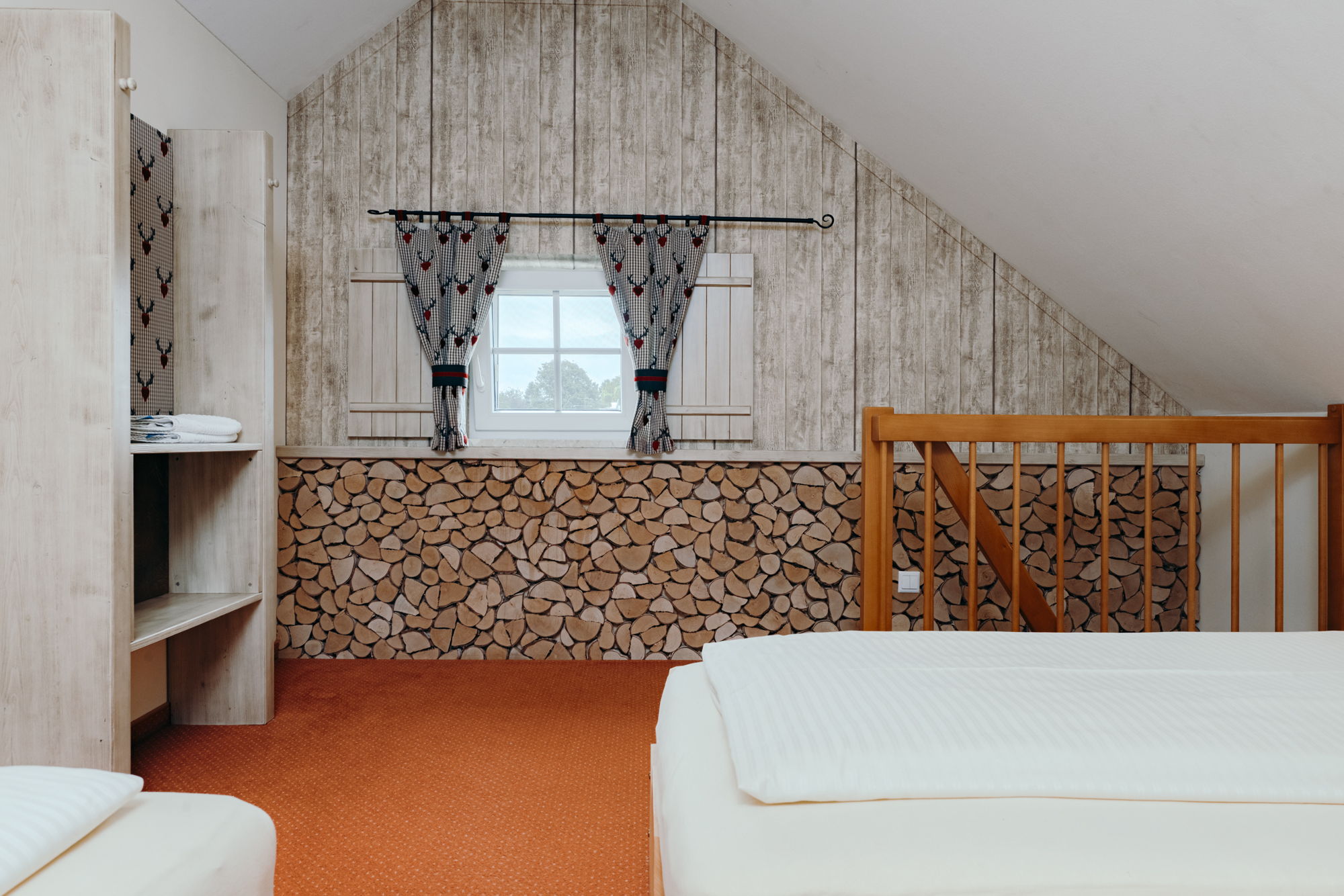 Ein Schlafzimmer mit Dachschräge, Treppenaufgang, weiß bezogenen Betten, im Hintergrund eine Wand mit Stein und Holz.