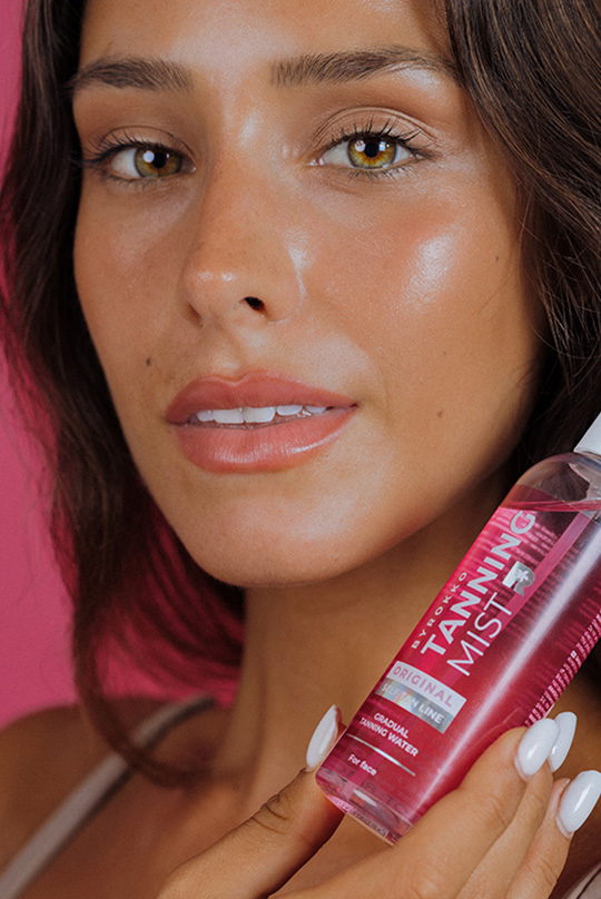 Nahaufnahme einer zufriedenen Kundin mit strahlender Selbstbräune, die eine Flasche ByRokko Strawberry Tanning Mist–Selbstbräunungsspray hält, um die Wirksamkeit und den ansprechenden Duft des Produkts zu verdeutlichen.