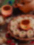 Festive Indian sweets shaped like small pumpkins, coated in coconut, arranged on a terracotta plate with decorative clay pots, cashews, rose petals, and a rustic clay teapot in the background.