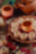 Festive Indian sweets shaped like small pumpkins, coated in coconut, arranged on a terracotta plate with decorative clay pots, cashews, rose petals, and a rustic clay teapot in the background.
