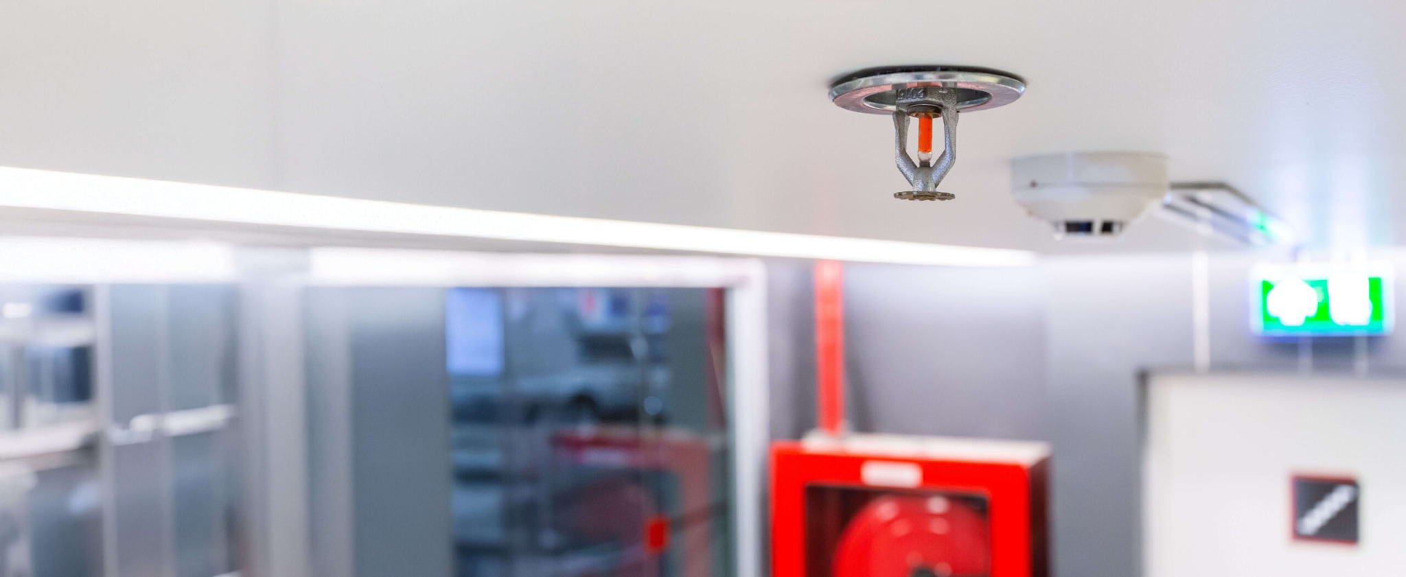 Close-up of a ceiling-mounted fire sprinkler in a modern building. A fire alarm and emergency exit sign are visible in the background, along with a red fire hose cabinet. The setting appears to be a safety-equipped indoor environment.