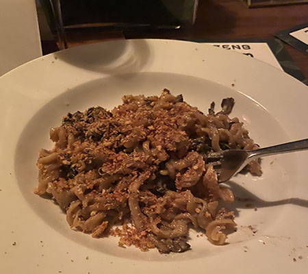 A closeup rotini pasta, seasonal mushrooms, and truffles, garnished with a breadcrumb crumble on a white plate with a fork.