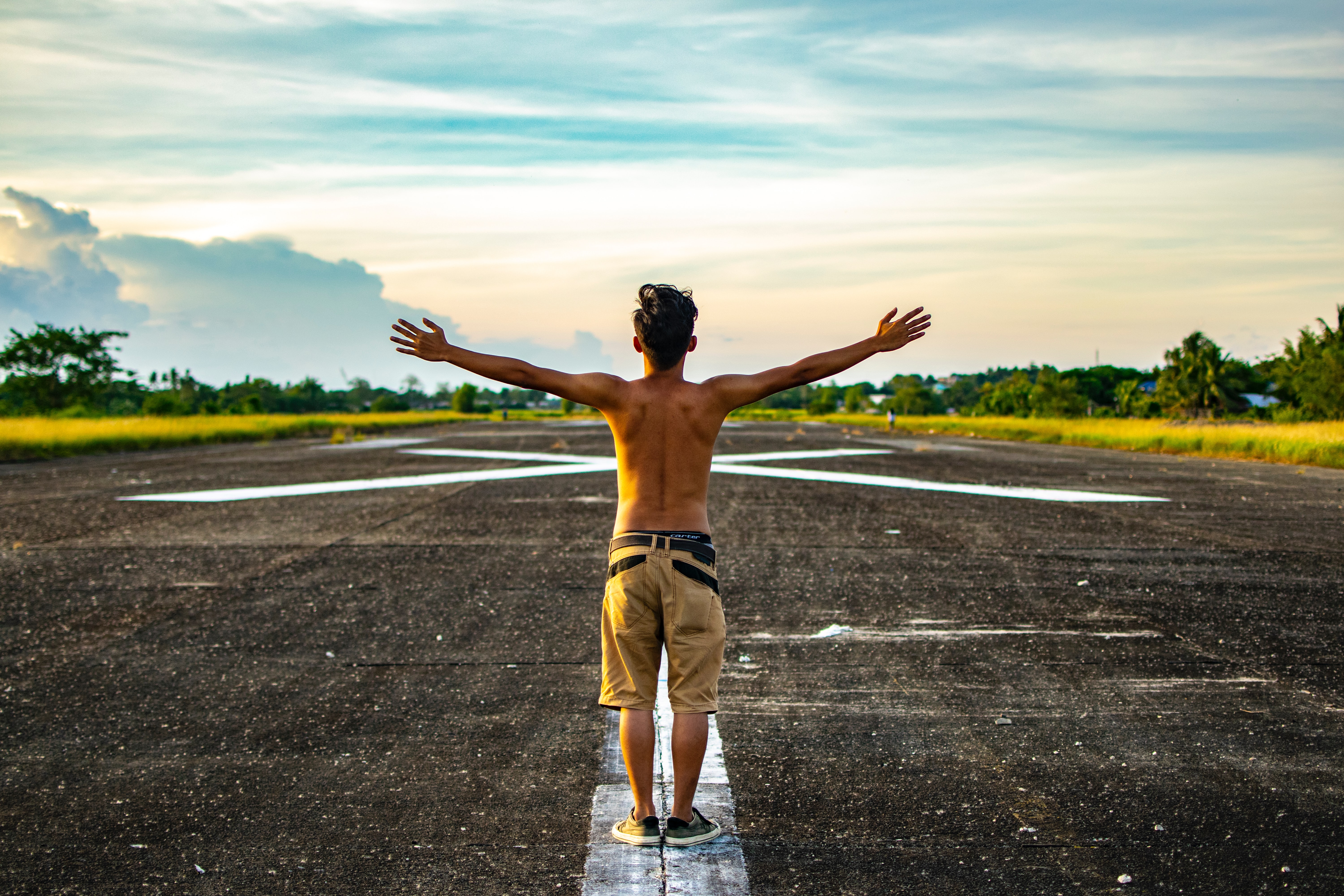 A shirtless guy stands in the middle of a white line on pavement with his arms up in the air.
