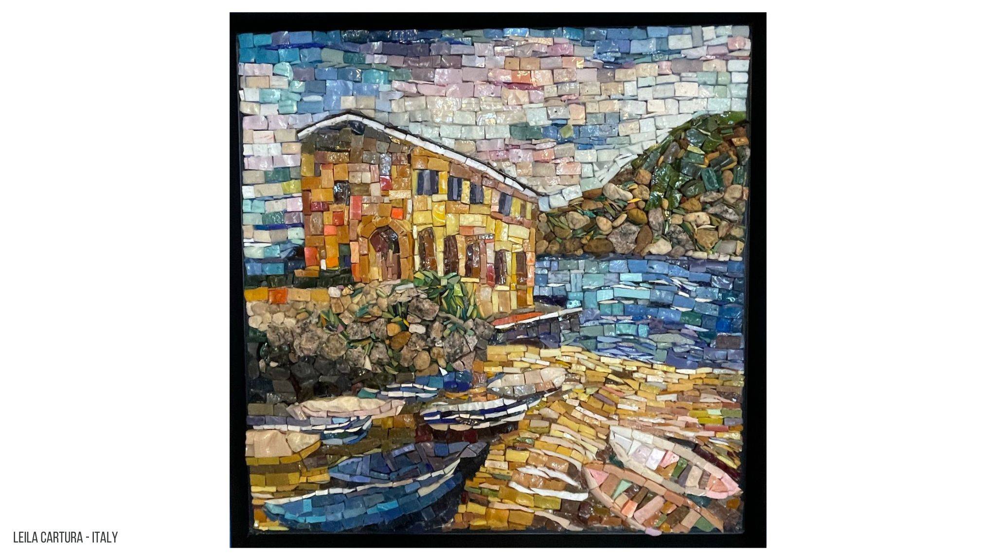 Een mozaiek van een gebouwtje aan een kleine haven met bootjes in het water gemaakt met Smalti. Op de achtergrond is een berg te zien die gemaakt is met stenen.