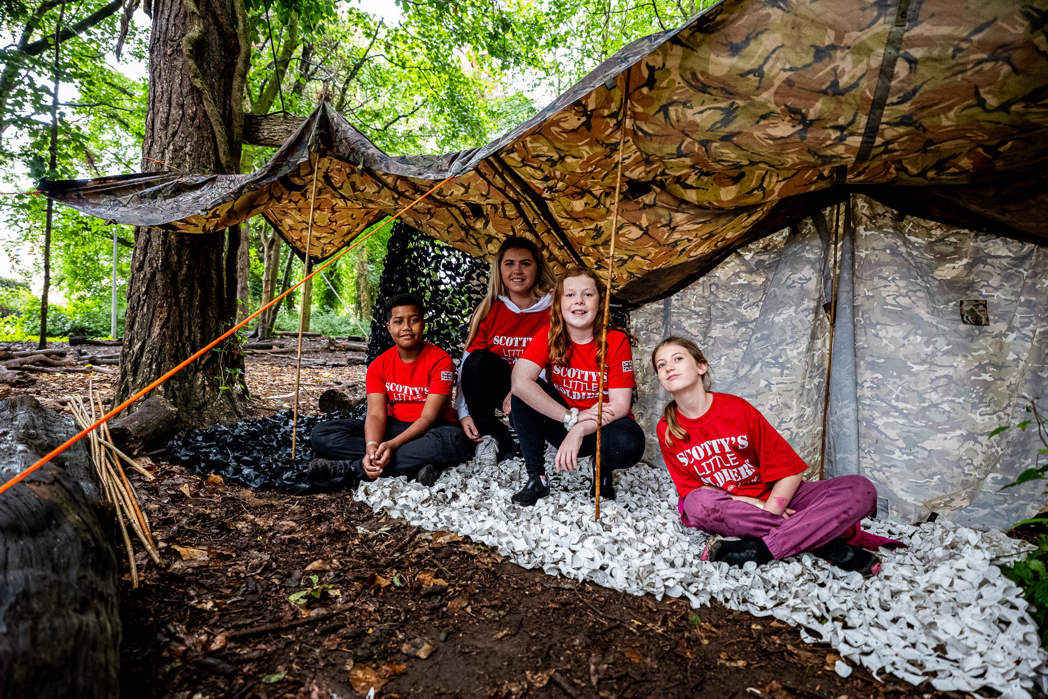 Four children wearing red Scotts Little Soldiers shirts sit and kneel under a camo tarp shelter in a wooded area, surrounded by trees and ground covered in leaves and white wood shavings.
