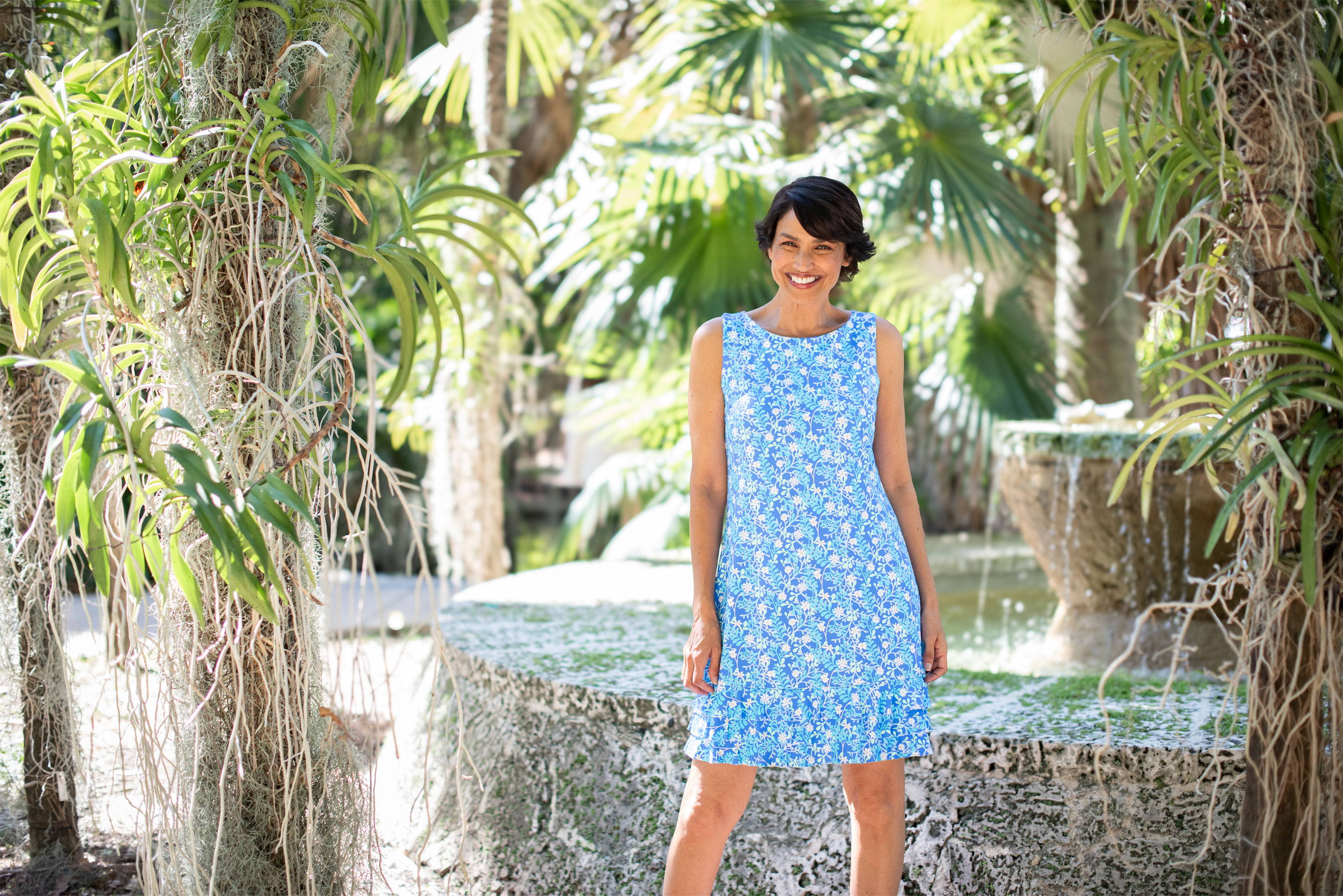 A woman wears a blue dress standing next to an outdoor fountain.