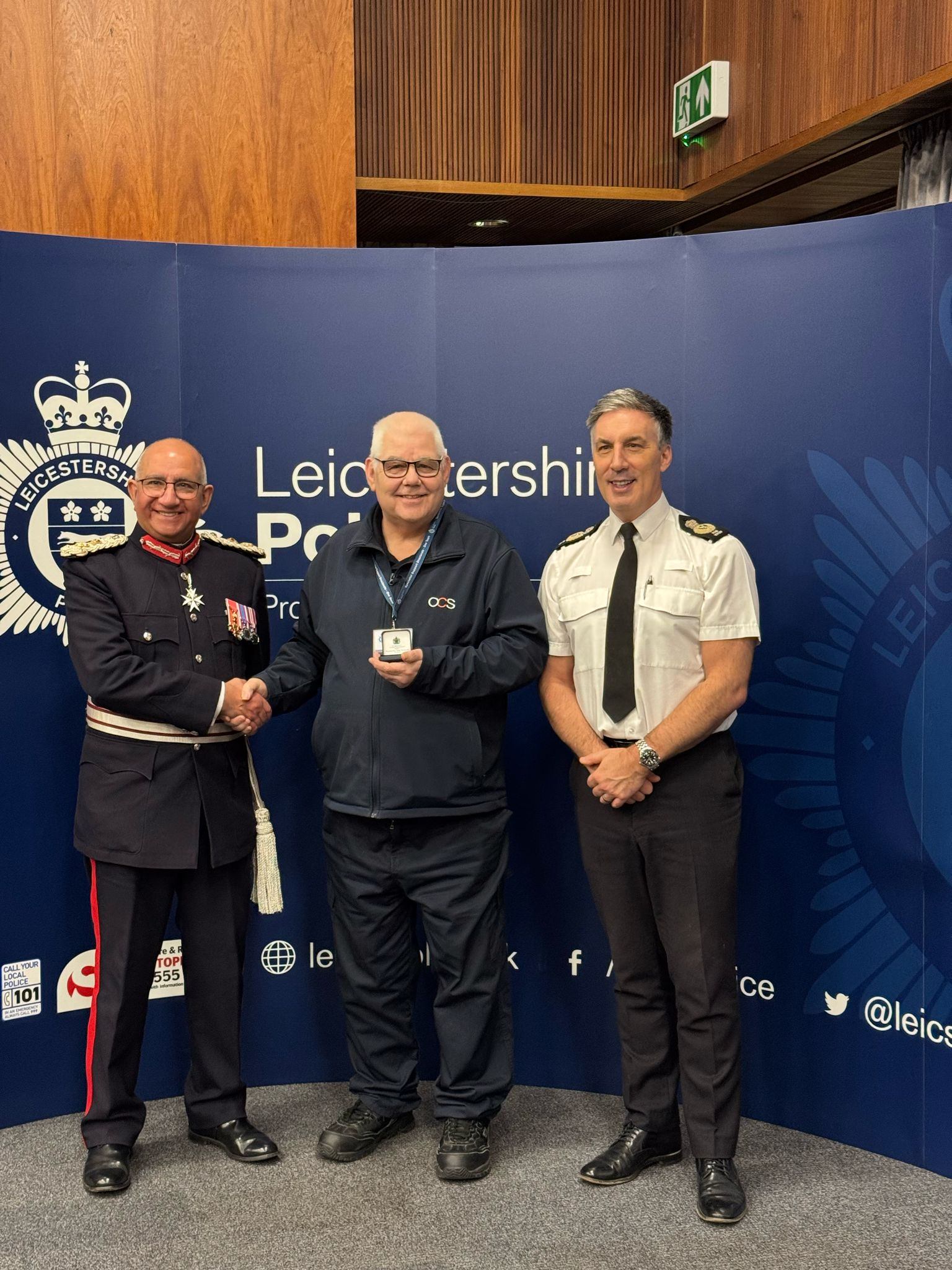 Three men are smiling and posing for a photo at an event, standing in front of a Leicestershire Police backdrop. One man is in a formal uniform with a sash, one is in a suit, and one is in a police uniform. The man in the suit holds a small box.