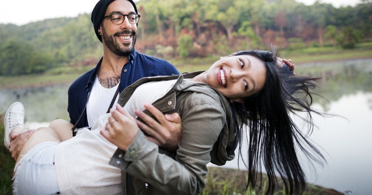 An attractive man and woman are next to a lake, the man is carrying the woman in his arms while she smiles and looks away.