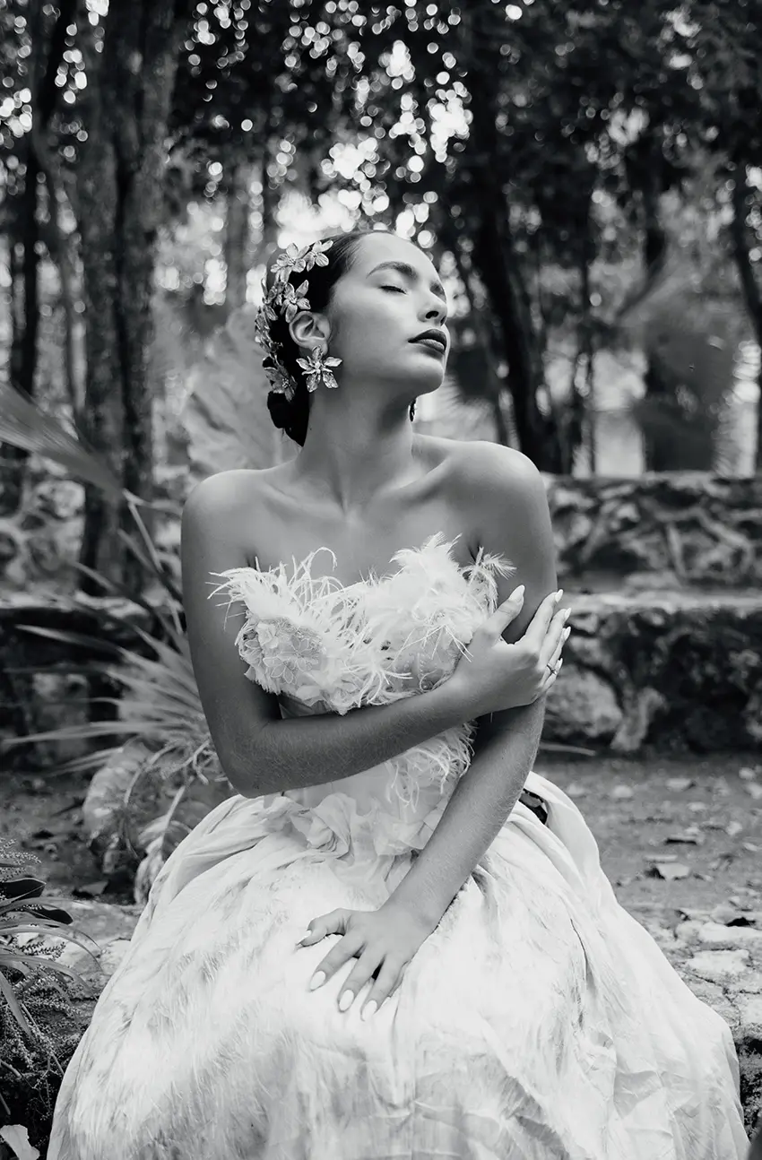 Une mariée dans une robe blanche avec des plumes, assise sur des rochers au Cenote R. La photographie est en noir et blanc, ce qui ajoute une touche artistique à la scène.