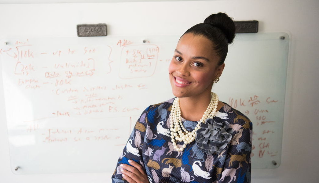 An attractive black woman with her arms crossed, in front of a whiteboard covered in writing. She has her hair in a bun and a cat long sleeve.