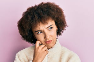 An attractive woman with large curls and puffy sweater looks pensive to the side. In front of a plain background.