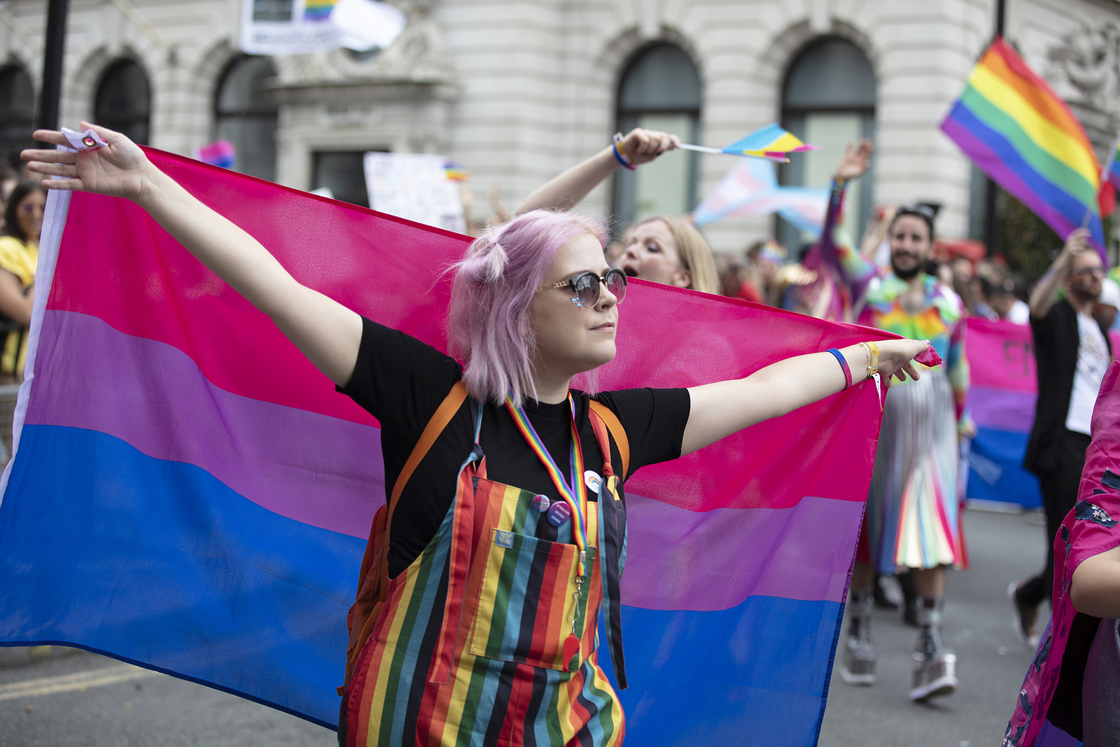 A woman with purple hair and rainbow suspenders holds the bi flat with her arms stretched out.