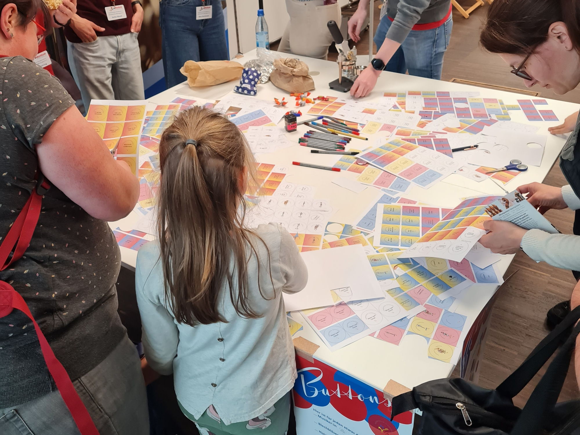 Auf einem Tisch liegen viele bunte Ausdrucke. Erwachsene und Kinder schneiden diese aus und basteln daraus Buttons. 