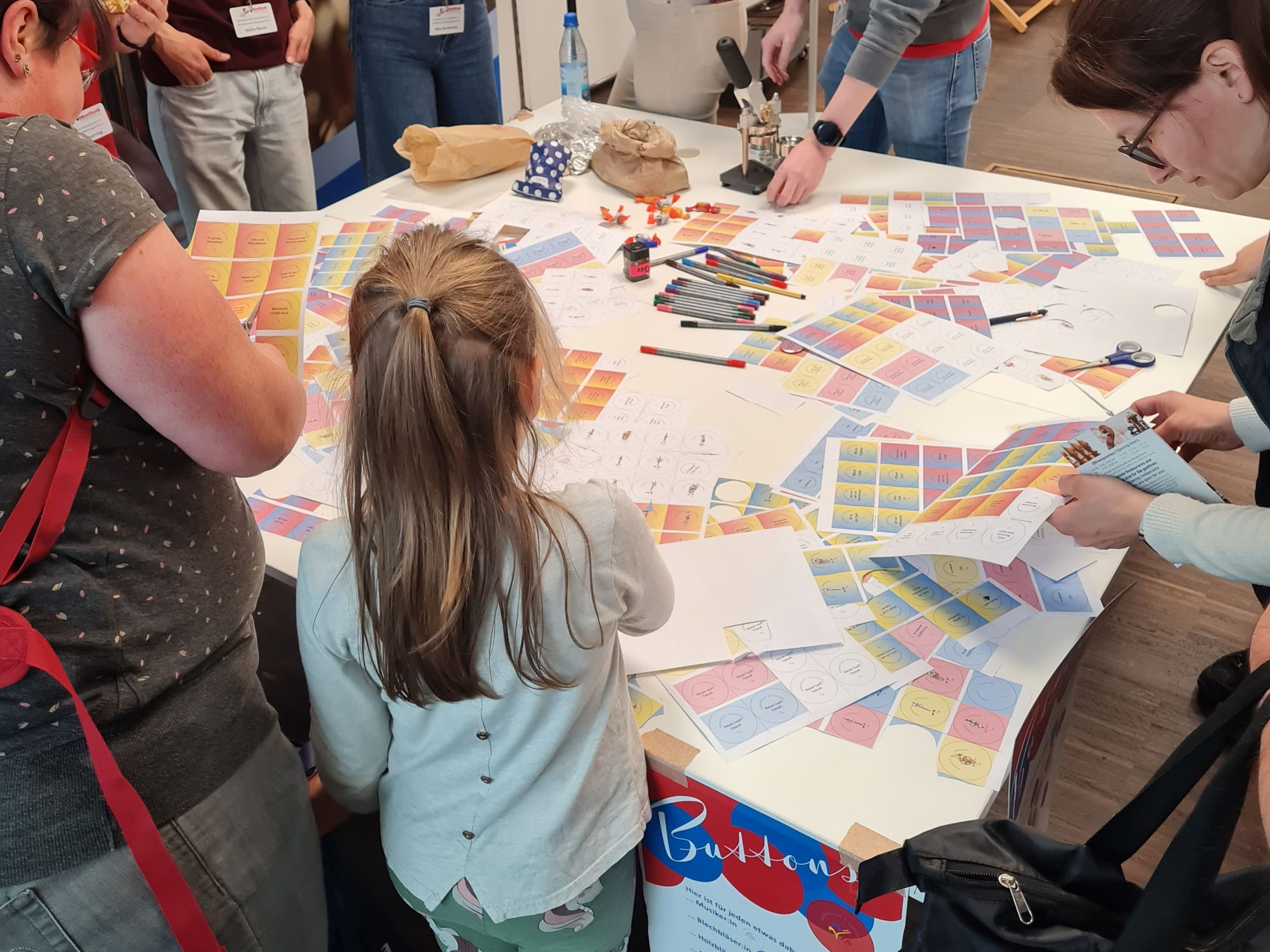 Auf einem Tisch liegen viele bunte Ausdrucke. Erwachsene und Kinder schneiden diese aus und basteln daraus Buttons. 