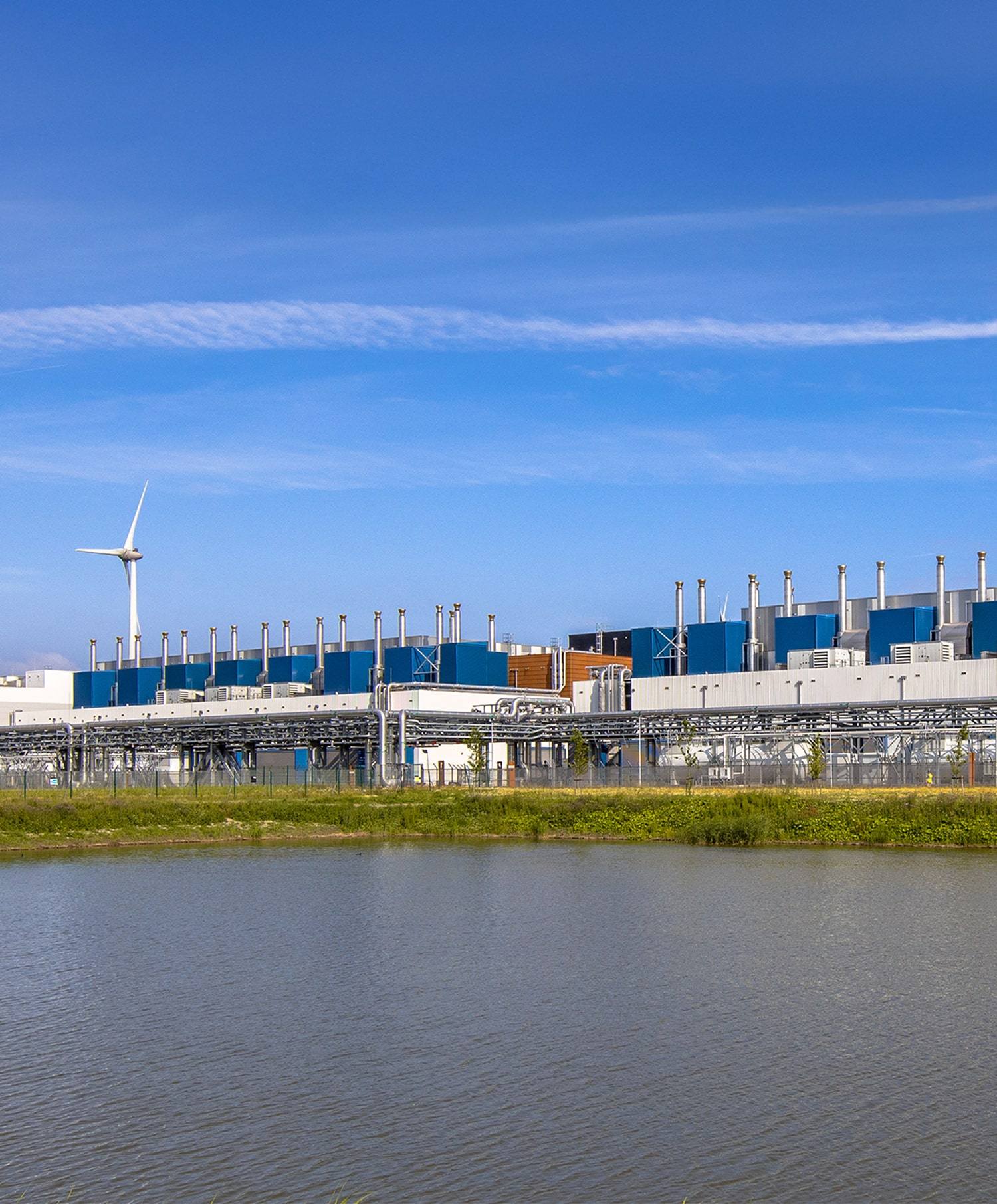 A modern industrial facility with blue and gray buildings and many smokestacks stands behind a body of water, with a wind turbine visible on the left under a clear blue sky.