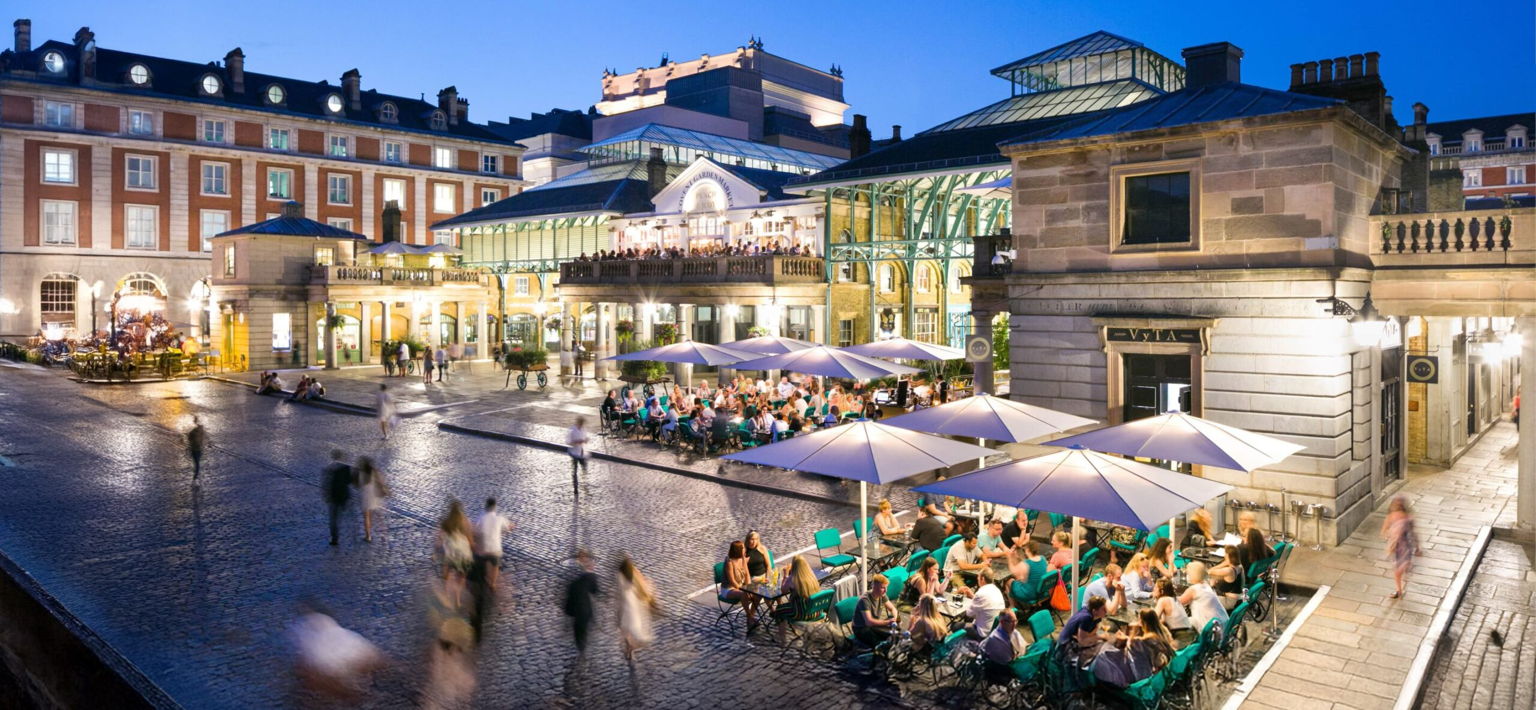 People sit at outdoor tables under large umbrellas in a lively plaza at dusk, with historic buildings and glass-roofed structures lit up in the background. The scene has a vibrant, welcoming atmosphere.