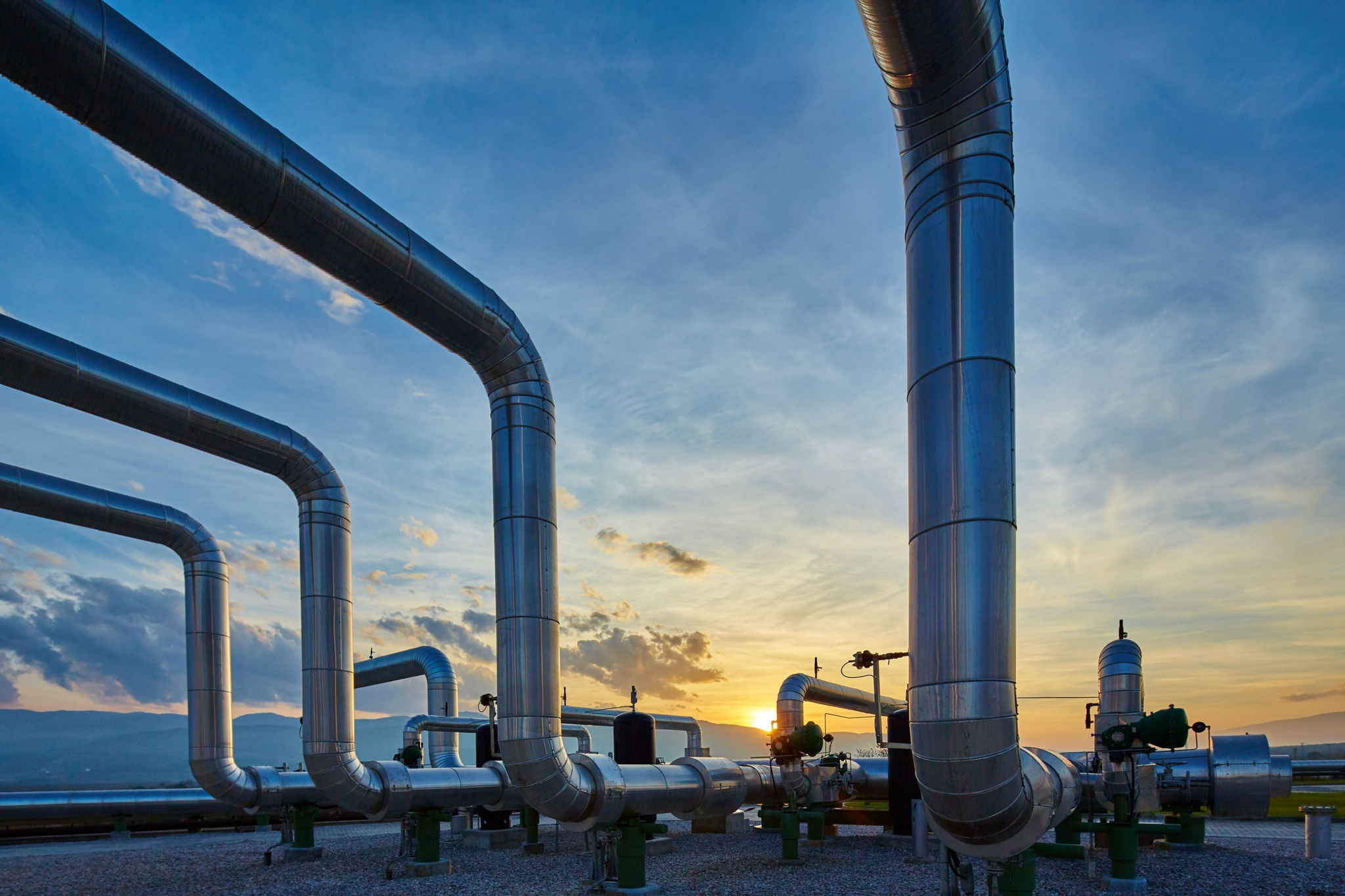 Large metal pipelines stretch across an outdoor industrial site at sunset, with a blue and orange sky in the background and some small mountains or hills on the horizon.