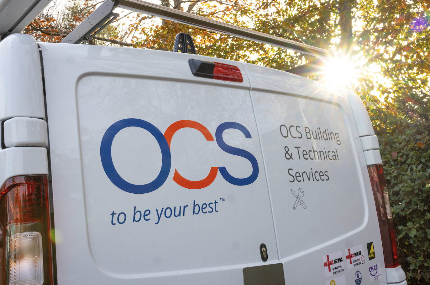 A white van with the OCS logo and the text to be your best™ and OCS Building & Technical Services on its back door, parked outdoors with sunlight shining through trees in the background.