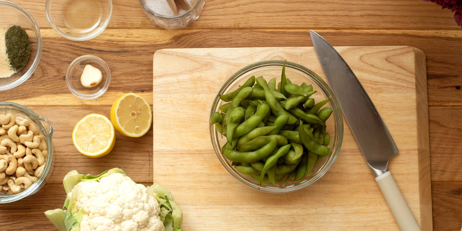 Bowl of edamame on cutting board surrounded by ingredients and a cutting knife.