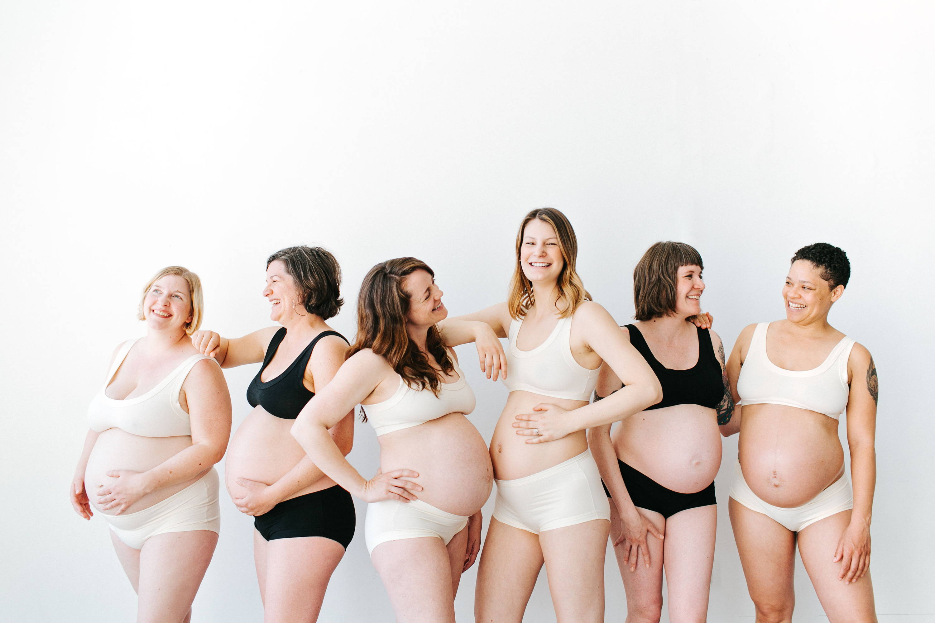 Six pregnant Women smiling at eachother wearing Thunderpants Organic Cotton Hipster and Original underwear with Bralettes in both plain Black and Vanilla colors.