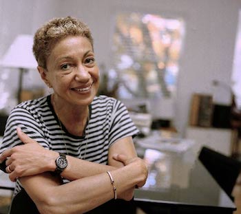 An older June Jordan sitting near a table crossing her arms and smiling warmly.