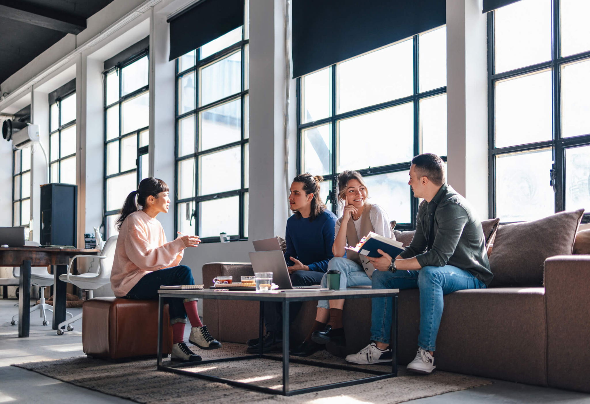 Four people sit on couches in a modern, sunlit office designed through our workplace services, engaged in discussion. Two laptops and notebooks are on the table. Large windows and open space create a relaxed atmosphere. One person gestures while others listen and smile.