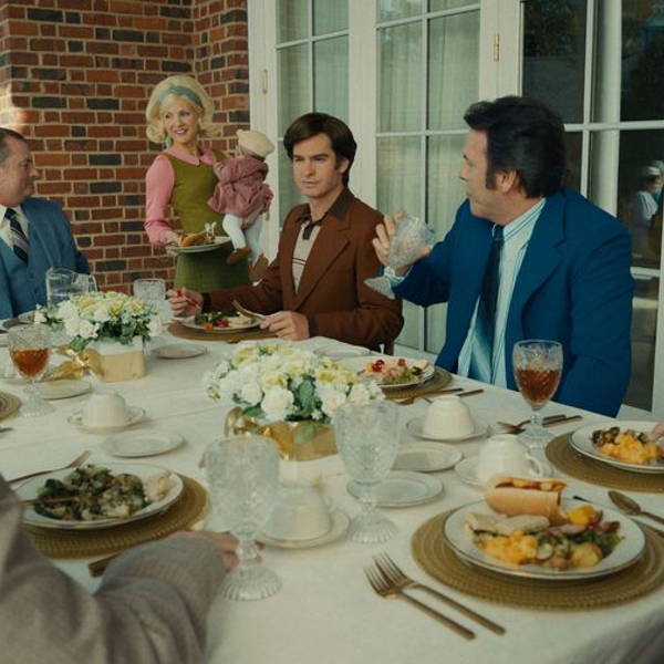 Men in suits eat at a table outside, interrupted by Tammy Faye who stands holding a child.