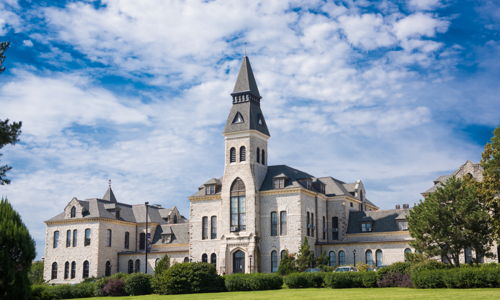 Historic building with tall spire and intricate stonework, set against a blue sky, surrounded by lush greenery.