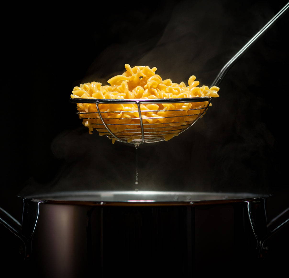 Rotini pasta in a strainer being pulled out of a black pot on a dark background with dramatic lighting