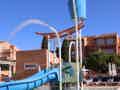 Eine blaue Wasserrutsche mit Kippeimer im Kinderbereich neben einem Pool mit Liegestühlen.