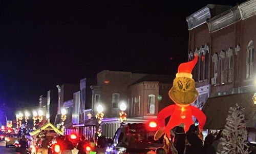 Festive nighttime parade scene with decorated vehicles, balloons, and a Grinch inflatable, evoking joy and community spirit.