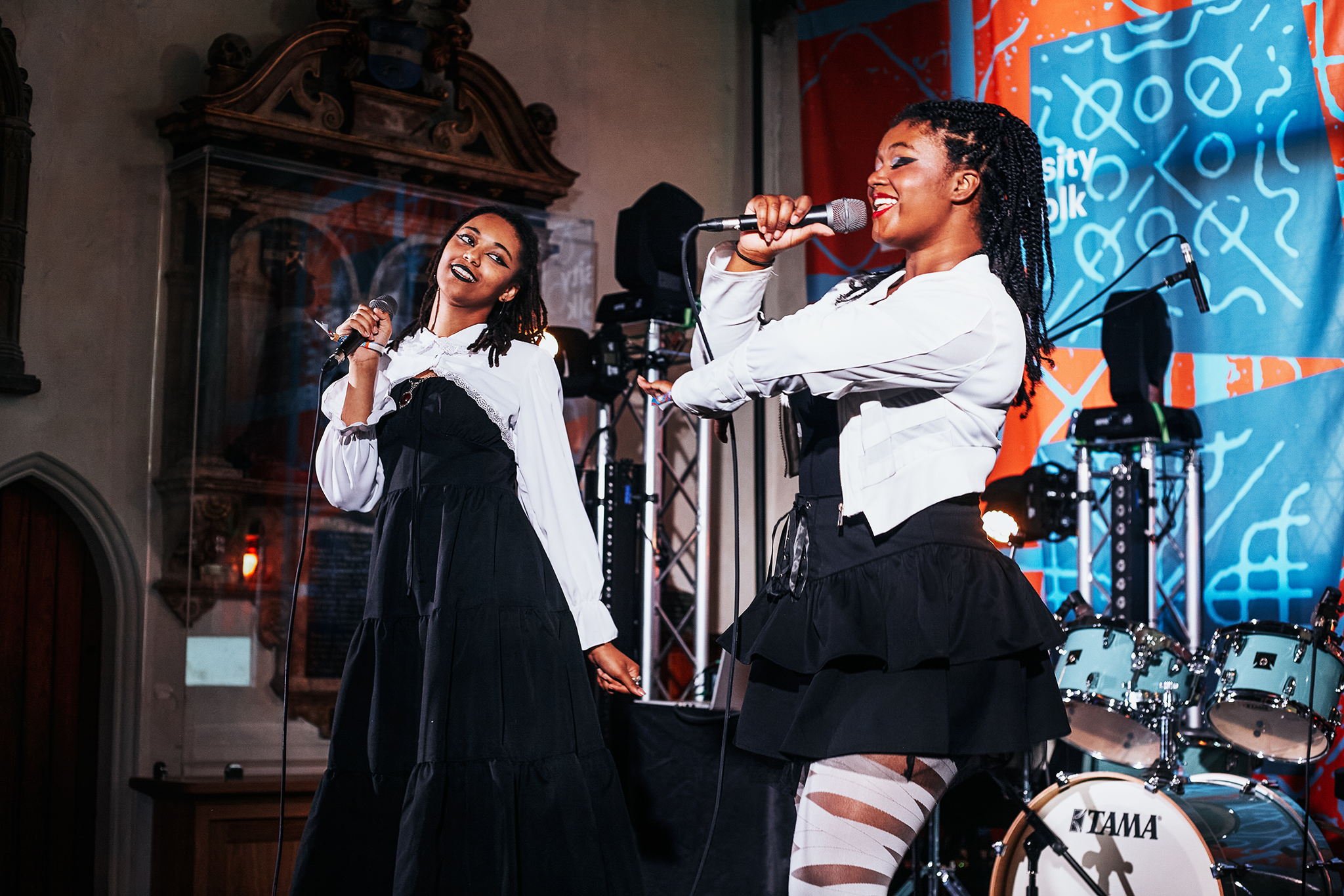 Two women in black and white outfits sing into microphones on stage. They smile and pose energetically, with stage lights, musical instruments, and colorful banners in the background.