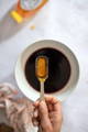A hand holds a measuring spoon of honey above a white bowl filled with a dark liquid, with a honey bottle and cloth nearby on a light surface.