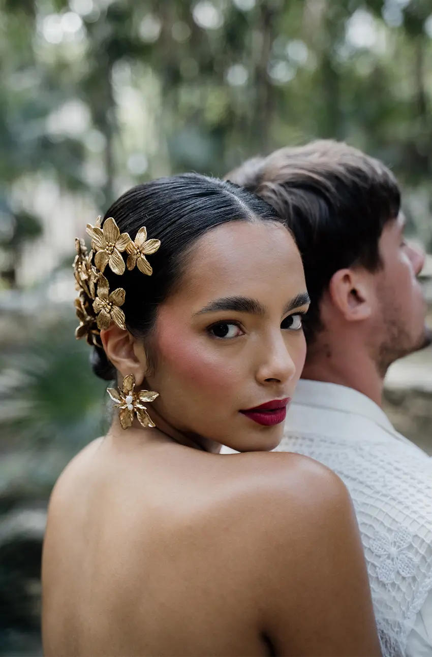 Une belle femme mexicaine aux cheveux noirs coiffés en chignon porte des boucles d'oreilles blanches et se tient au milieu de la luxuriante verdure des ruines anciennes de Tulum.