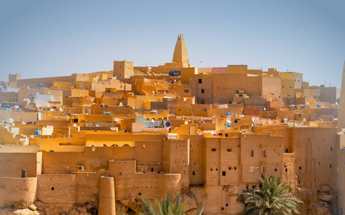 A town on a hillside, with matching tan buildings & rooftops, all getting higher into the clear blue sky. With trees at the bottom. 